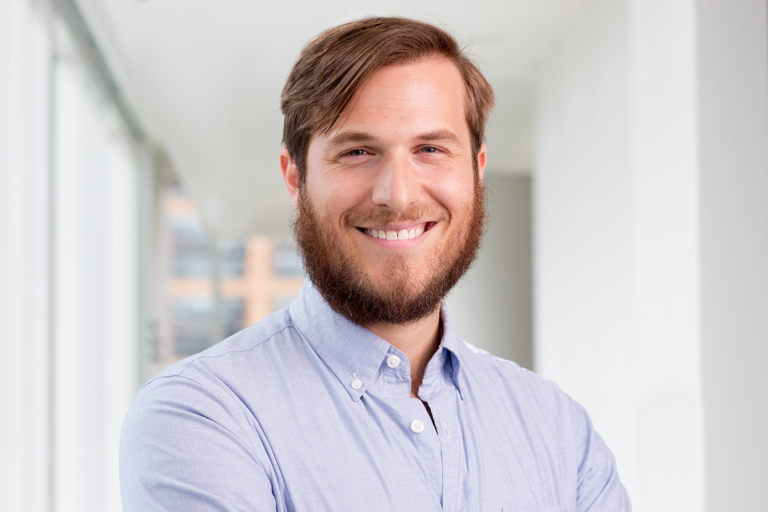 A white man with brown hair, beard, and mustache smiles facing the camera. He wears a light blue button-down.